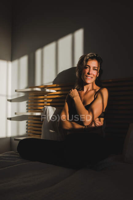 Стрілянина чуттєвої красивої жінки, яка посміхається між світлом і тінями в нижній білизні на ліжку, дивлячись на камеру — стокове фото