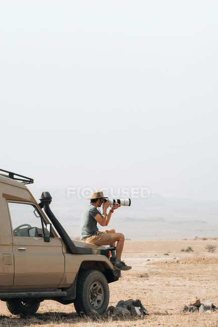 Вид збоку чоловіка, який подорожує фотографом, сидить на аутсордері і фотографується на камеру з телеоб'єктивом під час сафарі в савані влітку — стокове фото