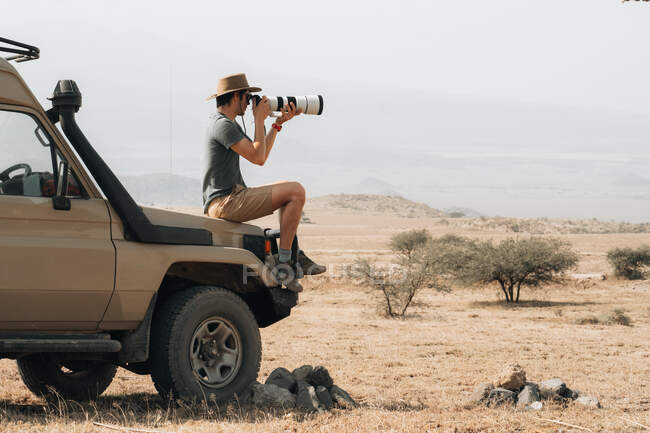 Vista lateral del fotógrafo viajero masculino sentado en offroader y tomando fotos en cámara con teleobjetivo durante el safari en Savannah en verano - foto de stock