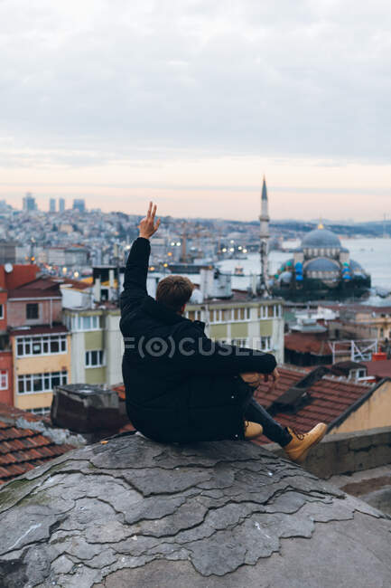 На тлі анонімного чоловічого туристичного жесту V знак сидячи на скелі проти житлового району з мечеттю та хмарним заходом сонця в місті Туреччина. — стокове фото