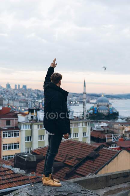 Back view of unrecognizable man standing with arm up on shabby rock edge against residential district and cloudy sundown sky with flying bird in evening in Turkey — Stock Photo