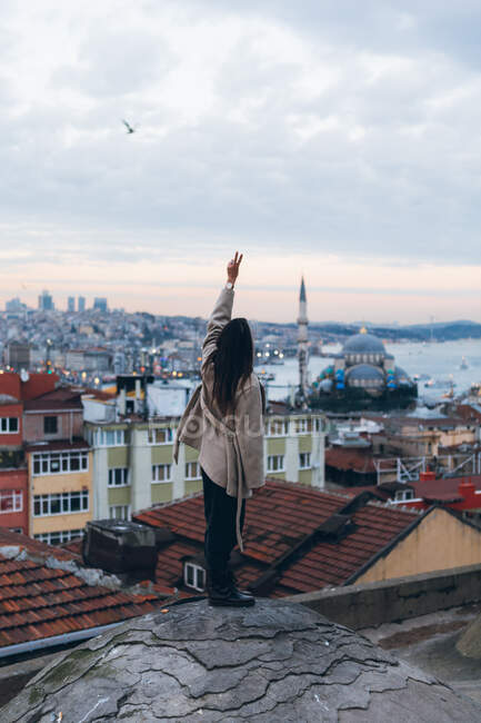 Visão traseira da fêmea na moda em pé na borda da rocha rasgada contra o bairro residencial e céu nublado ao pôr do sol à noite na Turquia — Fotografia de Stock