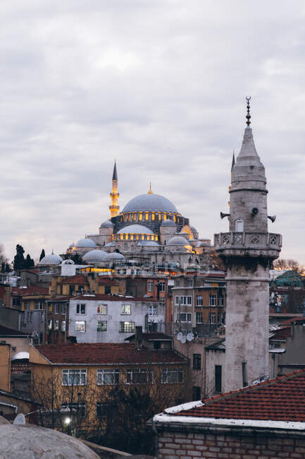 Вежа похилого віку, розташована серед обшарпаних будинків проти освітленої мечеті та хмарного неба на вулицях міста ввечері в Туреччині. — стокове фото