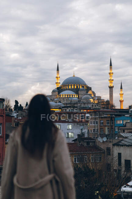 Back view of anonymous female tourist standing in residential city district and admiring illuminated mosque against cloudy sky in evening in Turkey — Stock Photo