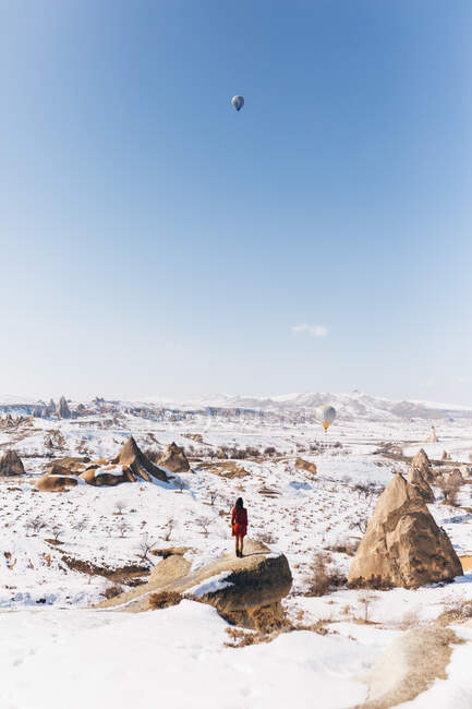 Полностью неузнаваемая женщина-туристка, стоящая на камне и любующаяся воздушными шарами, летающими в безоблачном голубом небе над снежной горной местностью в солнечный зимний день в Каппадокии, Турция — стоковое фото
