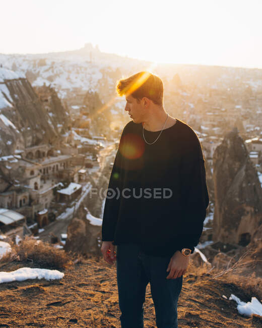 Jovem macho em roupa casual olhando para longe enquanto estava de pé contra o antigo assentamento de Uchisar coberto de neve e céu do pôr do sol na noite de inverno na Capadócia, Turquia — Fotografia de Stock