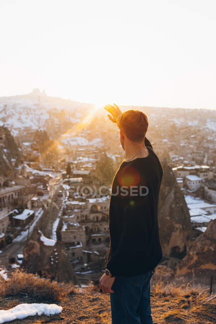 Seitenansicht eines nicht wiederzuerkennenden jungen Mannes in lässigem Outfit, der wegschaut, während er vor der alten Uchisar-Siedlung steht, die im Winterabend in Kappadokien, Türkei, mit Schnee und Sonnenuntergang bedeckt ist. — Stockfoto