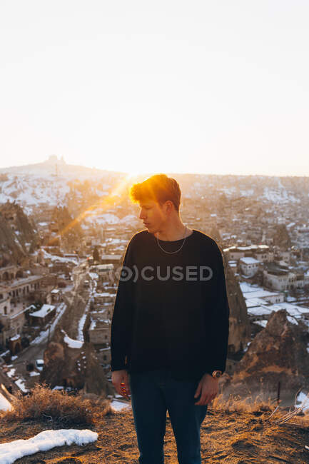 Young male in casual outfit looking away while standing against old Uchisar settlement covered with snow and sunset sky in winter evening in Cappadocia, Turkey — Stock Photo