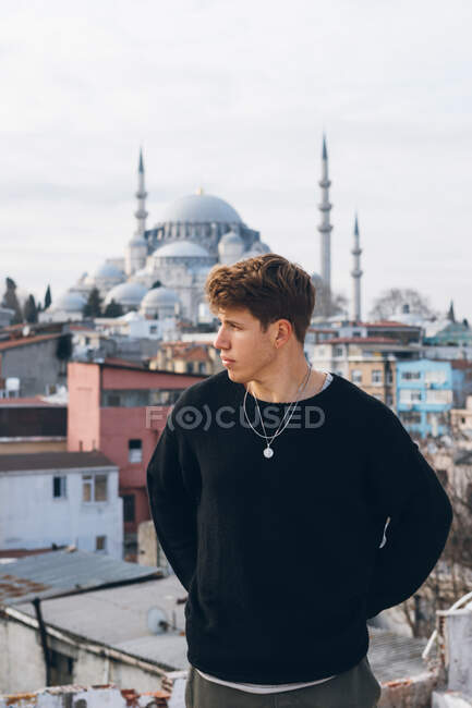 Jovem moderno em roupa casual olhando para longe, enquanto em pé contra edifícios pobres de bairro residencial e mesquita tradicional em dia nublado na cidade na Turquia — Fotografia de Stock
