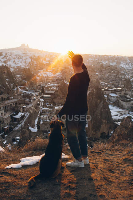 Back view of anonymous young man petting dog and covering eyes from bright sunset sun while admiring aged Uchisar settlement in evening in Cappadocia, Turkey — Stock Photo