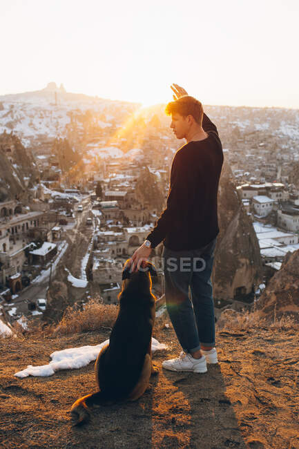 Side view of young man petting dog and covering eyes from bright sunset sun while admiring aged Uchisar settlement in evening in Cappadocia, Turkey — Stock Photo