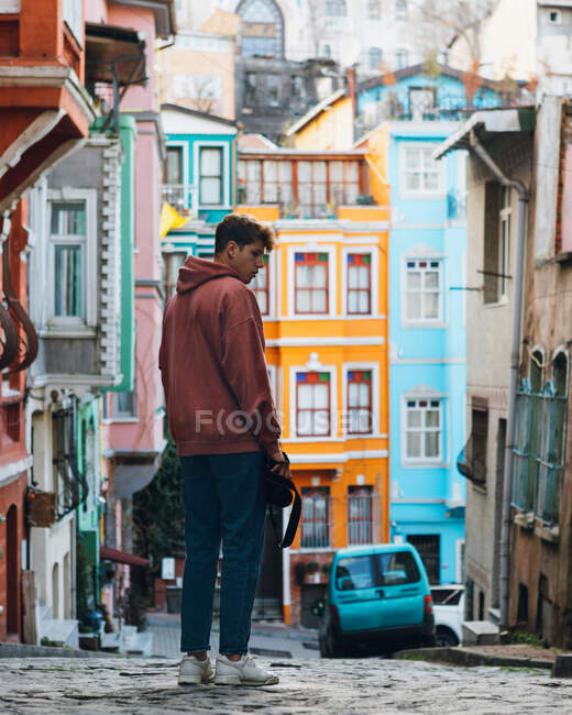 Visão traseira do jovem com câmera de foto em pé na estrada pavimentada perto de casas coloridas e carro azul na rua da cidade na Turquia — Fotografia de Stock