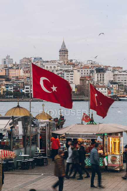 Moderne Menschen gehen an einem trüben Tag in der Stadt in der Türkei in der Nähe von Verkaufsständen und Nationalflaggen auf einem Damm am Fluss — Stockfoto
