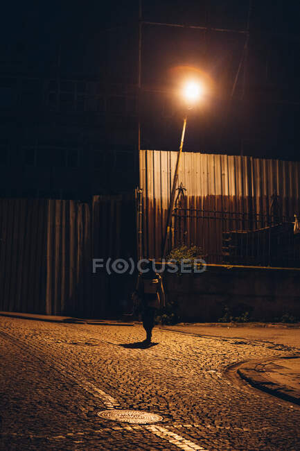 Silueta de hombre irreconocible caminando solo a lo largo de la calle bajo la iluminación de farola brillante en la ciudad - foto de stock