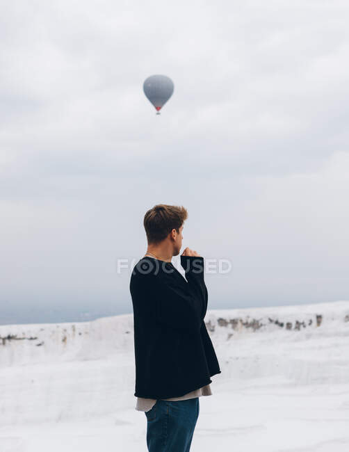 Путешествуя неузнаваемым человеком в повседневной одежде, смотрящим в сторону, стоя на белом холме минерального образования против сельской местности на горизонте и воздушном шаре, летящем в сером небе в Турции — стоковое фото
