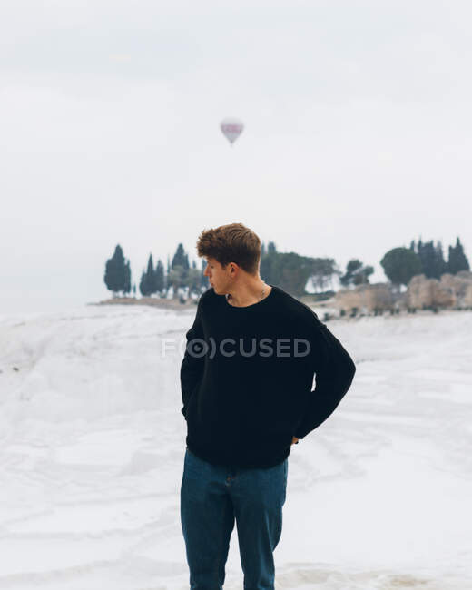 Путешествующий человек в повседневной одежде смотрит через плечо, стоя на белом холме минерального образования против сельской местности на горизонте и воздушный шар, летящий в сером небе в Турции — стоковое фото