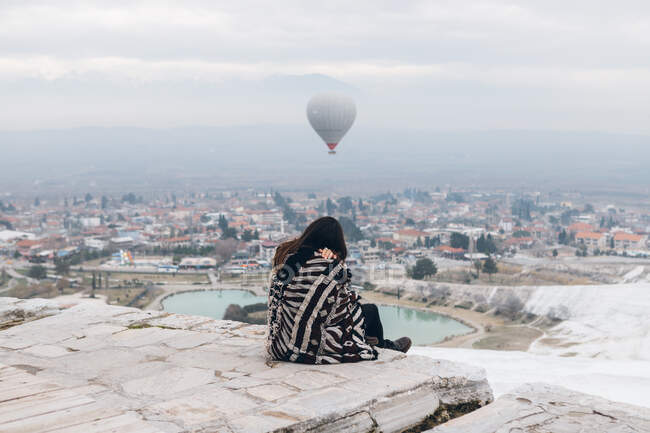 Back view of unrecognizable melancholic female in warm clothes sitting on stone slabs and embracing self while watching for big gray air balloon racing in cloudy sky over foggy city in overcast cool weather in Turkey — Stock Photo