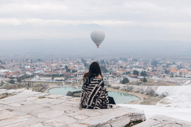 Погляд на нерозпізнану меланхолійну самицю в теплому одязі, яка сидить на кам'яних плитах і обіймає себе, спостерігаючи за бігом великого сірого повітряного балона в хмарному небі над туманним містом у спекотну погоду в Туреччині. — стокове фото