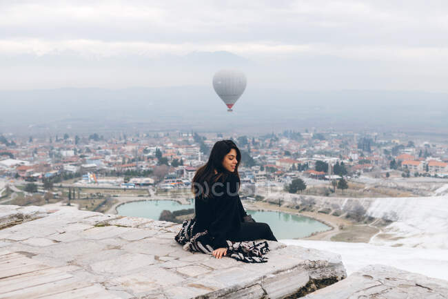 Melancholic female in warm clothes sitting with closed eyes on stone slabs and embracing self while watching for big gray air balloon racing in cloudy sky over foggy city in overcast cool weather in Turkey — Stock Photo