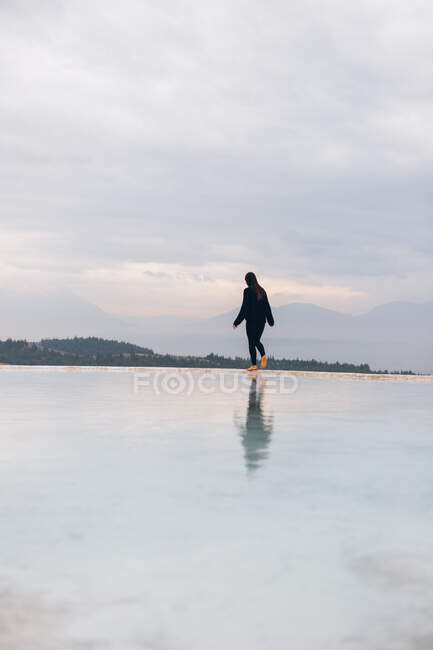 Back view of unrecognizable woman in vivid clothes with arms outstretched walking along coast of white mineral formation washing by clear water in Turkey — Stock Photo