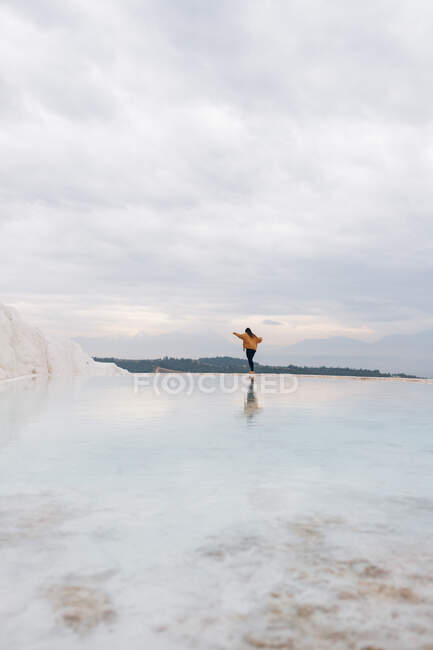 Обратный вид на неузнаваемую женщину в яркой одежде с протянутыми руками, идущую вдоль берега белого минерального образования, омываемую чистой водой в Турции — стоковое фото