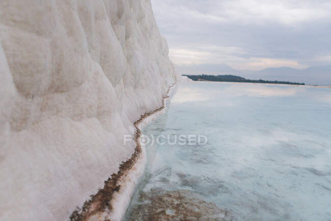 Formation de sel minéral sur le littoral se lavant par une eau claire et transparente contre les collines boisées brumeuses à l'horizon par temps couvert en Turquie — Photo de stock