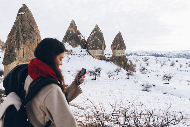 Side view of female traveler in warm clothes using smartphone while standing against snowy hills with famous pillars with sharp spear shaped peaks in national park in Turkey — Stock Photo