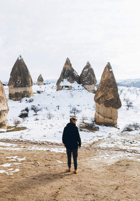 Rückansicht eines Mannes in dunkelwarmer Kleidung, der wegschaut, während er auf einem Feldweg gegen einen schneebedeckten Hügel mit berühmten Säulen mit scharfen, speerförmigen Gipfeln im Nationalpark in der Türkei steht — Stockfoto