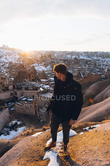 Von oben ein Mann in warmer Kleidung, der bei Sonnenuntergang in der Türkei auf einem verschneiten Hügel vor einer berühmten Kleinstadt mit alten Höhlenhäusern im Tal steht und über die Schulter schaut — Stockfoto