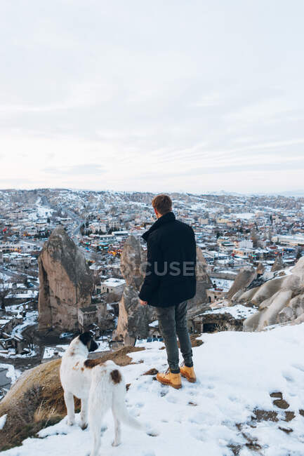 Rückansicht eines jungen, nicht wiederzuerkennenden Mannes in warmer Kleidung, der mit seinem treuen Hund auf einem Hügel vor kleinen alten Höhlenhäusern in einem verschneiten Tal in der Abenddämmerung in der Türkei steht — Stockfoto