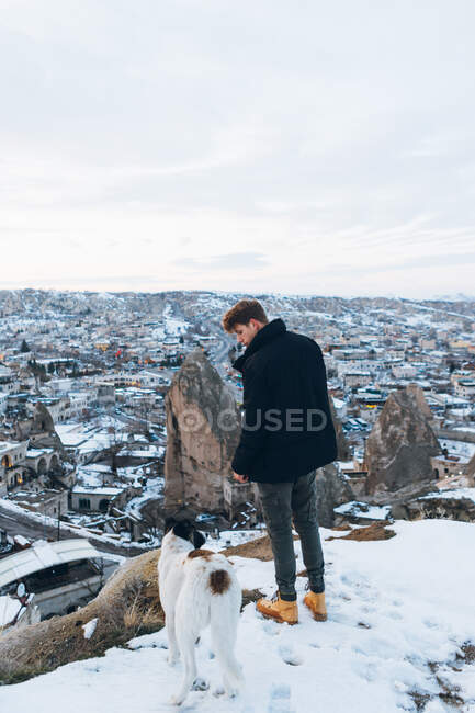 Vista posteriore di un giovane in abiti caldi in piedi con leale cane sulla collina contro piccole antiche case rupestri nella valle innevata al tramonto in Turchia — Foto stock