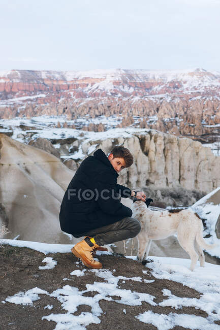 Young man squatted down in warm clothes caressing dog in snowy hill against misty mountains in overcast weather in Turkey — Stock Photo
