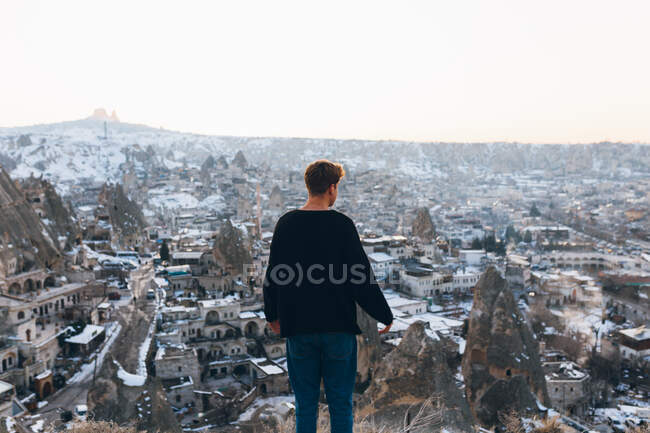 Back view of unrecognizable young man in warm clothes standing on snowy hill looking over famous small town with ancient cave houses in valley during sunset in Turkey — Stock Photo