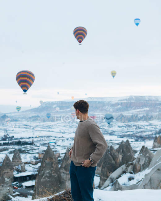 Seitenansicht eines nicht wiederzuerkennenden Mannes, der bei trübem Wetter in der Türkei vor ungewöhnlichen Steinsäulen und bunten Luftballons steht, die in den Himmel über nebligem, verschneitem Hochland rasen — Stockfoto