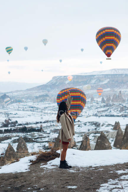 Vista lateral da fêmea em roupas quentes olhando para cima, enquanto em pé contra pilares de pedra incomuns e balões de ar coloridos correndo no céu sobre montanhas nebulosas nevadas em tempo nublado na Turquia — Fotografia de Stock