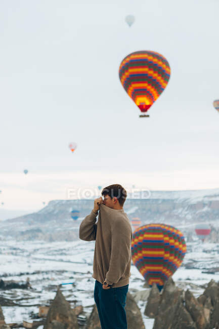 Vista lateral do homem olhando para a paisagem enquanto está de pé contra pilares de pedra incomuns e balões de ar coloridos correndo no céu sobre montanhas nebulosas nevadas em tempo nublado na Turquia — Fotografia de Stock