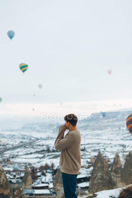 Vista lateral do homem irreconhecível olhando para a paisagem enquanto está de pé contra pilares de pedra incomuns e balões de ar coloridos correndo no céu sobre montanhas nebulosas nevadas em tempo nublado na Turquia — Fotografia de Stock