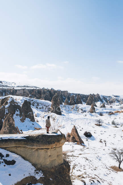 Сверху на камне стоит неузнаваемая туристка, любуясь удивительными снежными пейзажами на безоблачное голубое небо над снежной горной местностью в солнечный зимний день в Каппадокии, Турция — стоковое фото