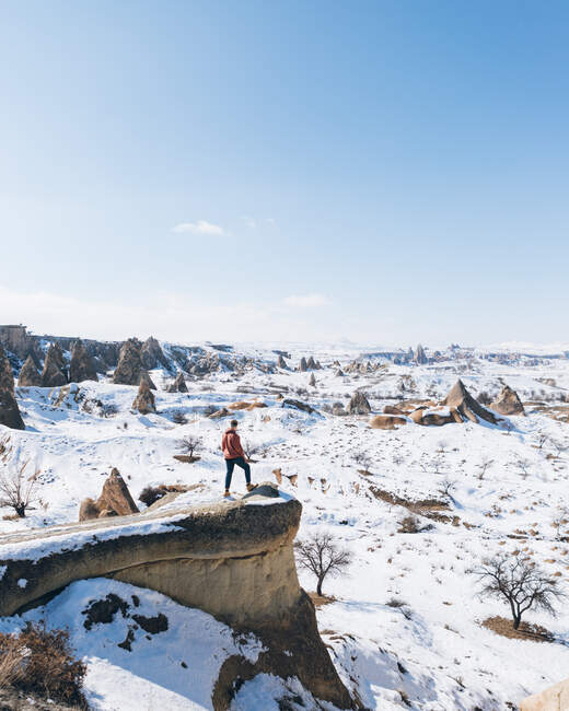 De acima de corpo inteiro turista homem irreconhecível em pé na pedra e admirando incríveis vistas de paisagem nevadas no céu azul sem nuvens sobre terreno montanhoso nevado no dia ensolarado de inverno na Capadócia, Turquia — Fotografia de Stock