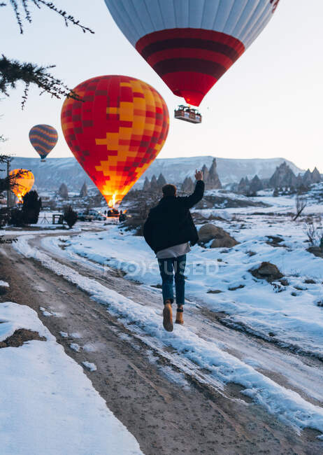 Back view of unrecognizable man in warm clothes with arm raised running to vivid colored air balloon flying up into gray sky over foggy snowy mountain ridge in Turkey — Stock Photo