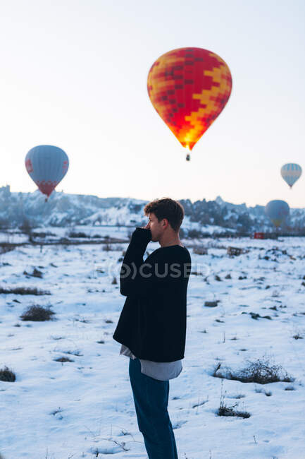 Side view of man in warm clothes looking away while standing with vivid colored air balloon flying up into gray sky over foggy snowy mountain ridge in Turkey — Stock Photo