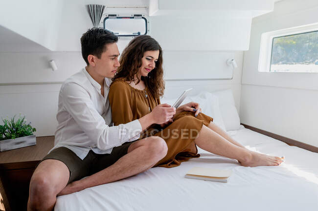 Vue latérale d'un couple amoureux se relaxant au lit en cabine de catamaran et tablette de navigation ensemble pendant le voyage en mer — Photo de stock