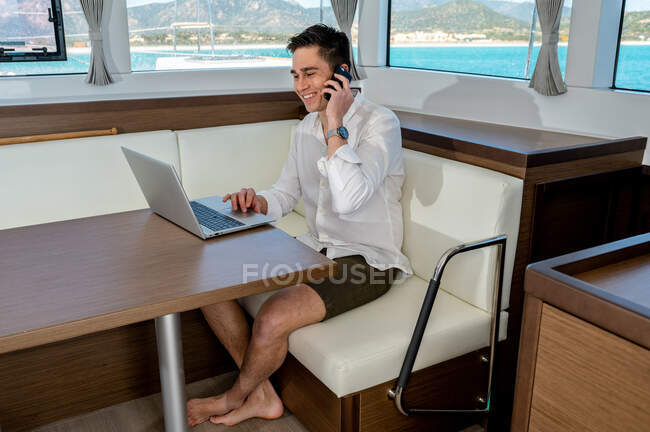 Vista lateral de alto ángulo del contenido freelancer masculino sentado en el cuento en la cabina del catamarán y trabajando mientras habla en el teléfono inteligente y el ordenador portátil de navegación - foto de stock