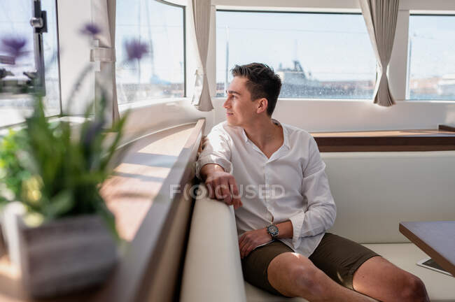 Délicieux mâle assis sur le canapé dans la cabine du catamaran et regardant par la fenêtre tout en profitant des vacances d'été en mer — Photo de stock