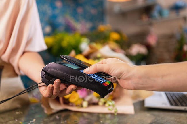 Ritaglia il cliente irriconoscibile effettuando il pagamento senza contatto con il telefono cellulare sul terminale POS nel negozio di fiori — Foto stock