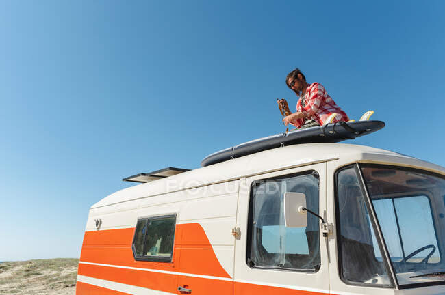 Низький кут молодого мандрівника чоловічої статі у повсякденному одязі та сонцезахисних окулярах фіксує серфінг на даху фургона проти безхмарного блакитного неба. — стокове фото
