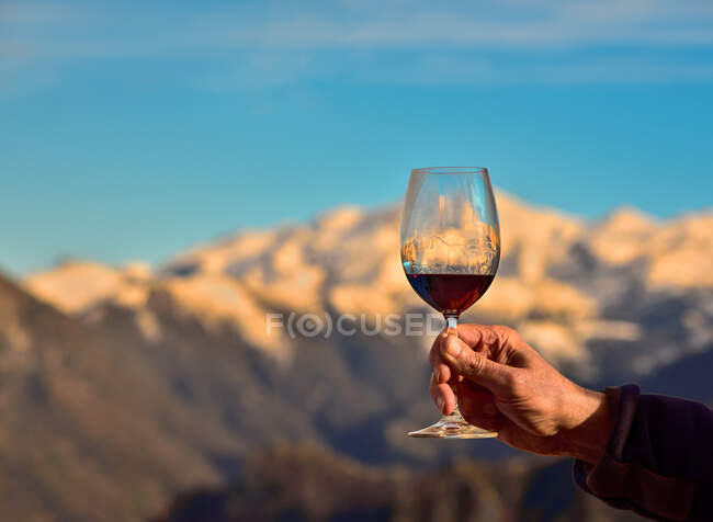 Ireconhecível pessoa madura propondo brinde com copo de vinho tinto no fundo borrado do cume da montanha e céu azul — Fotografia de Stock