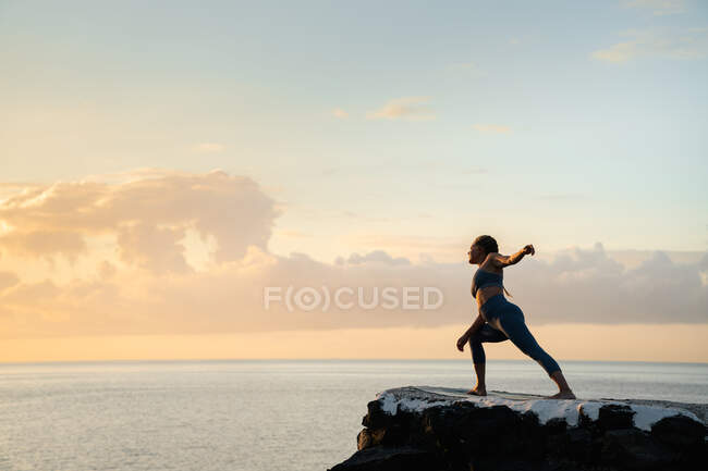 Seitenansicht von Yoga praktizierenden Frauen auf Utthita Parsvakonasana posieren auf Felsbrocken gegen den Ozean unter bewölktem Himmel bei Sonnenuntergang — Stockfoto