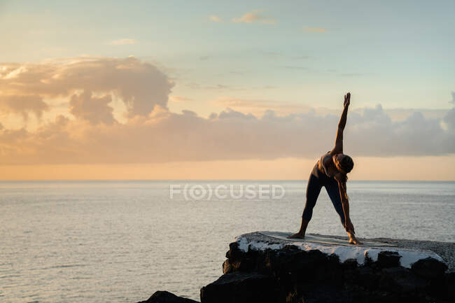 Unrecognizable female in sportswear standing in Trikonasana pose on yoga mat against rippled ocean under cloudy sky at sunset — Stock Photo