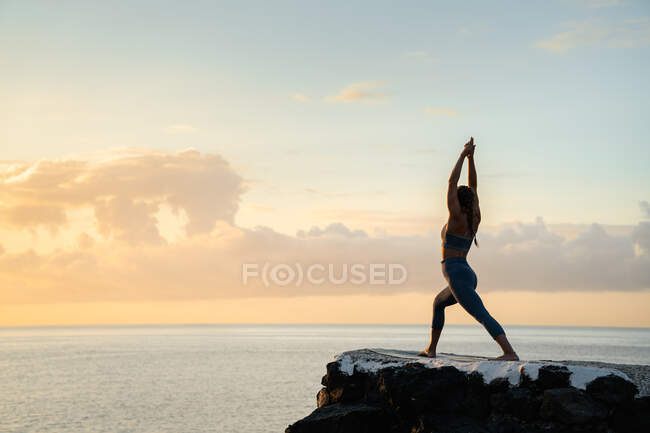 Side view of anonymous female practicing yoga with raised arms on boulders against ocean under cloudy sky at sundown — Stock Photo